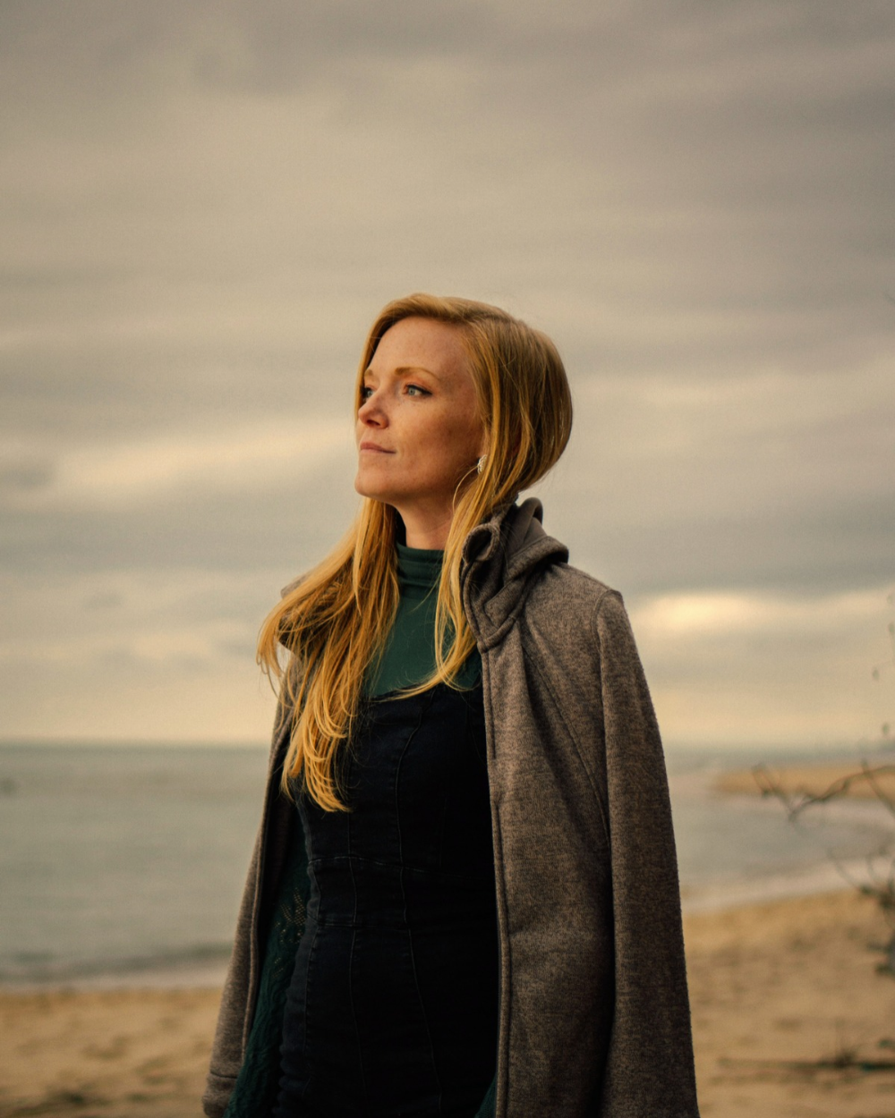 A woman with long blonde hair in a grey coat standing on an overcast beach