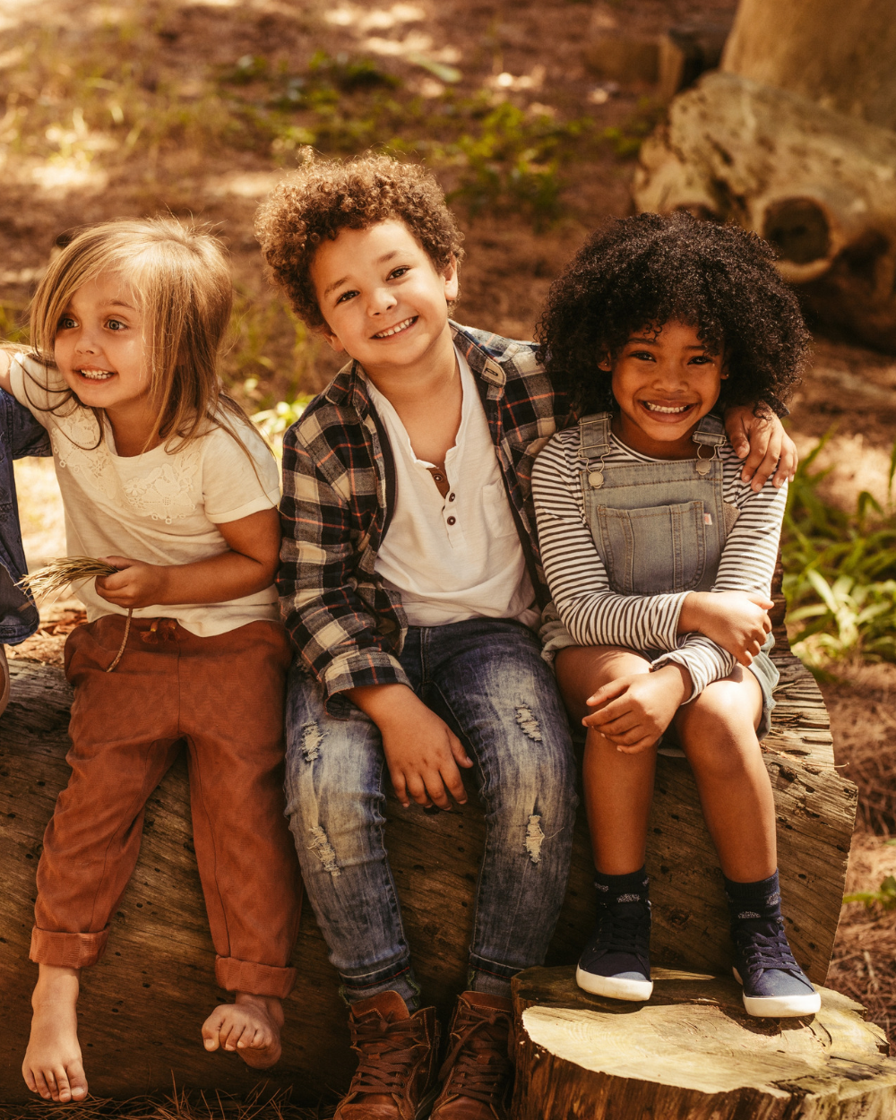 Three smiling children sitting together on a log in a sunlit forest